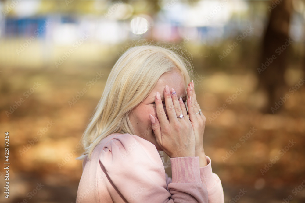 Smiling blond woman sitting in the park and looking at camera. Holding hands on face.