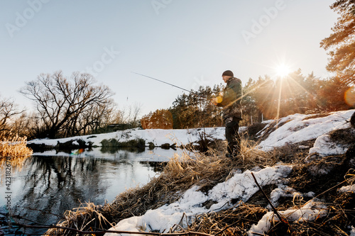 A fisherman with a fishing rod catches fish on the bank of a snow-covered river in early spring