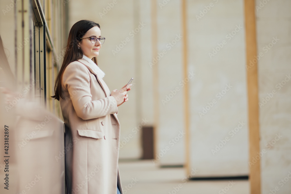 Portrait of a young business woman holding phone against panoramic windows and looking away and thinking. Portrait of a young business woman holding phone against panoramic windows and looking away and thinking.