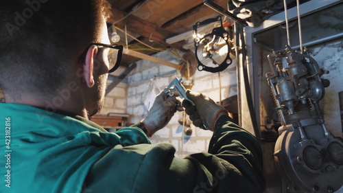 Male mechanic in protective glasses examines vehicle detail in garage. Male worker using tool for his work at workplace. Young repairman in uniform working in workshop. Close up Slow motion