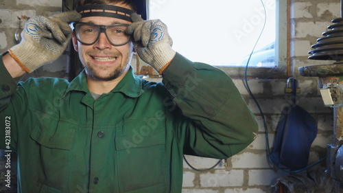 Young repairman taking off protective eyewear and showing grimaces into camera. Portrait of welder in uniform making funny faces and fooling around in garage. Mechanic with beard working in workshop © olehslepchenko