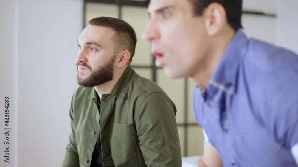 Young bearded Caucasian man watching TV with Middle Eastern friend indoors. Side view portrait of absorbed handsome mates enjoying weekend leisure at home. Lifestyle and friendship. Young bearded Caucasian man watching TV with Middle Eastern friend indoors. Side view portrait of absorbed handsome mates enjoying weekend leisure at home. Lifestyle and friendship.