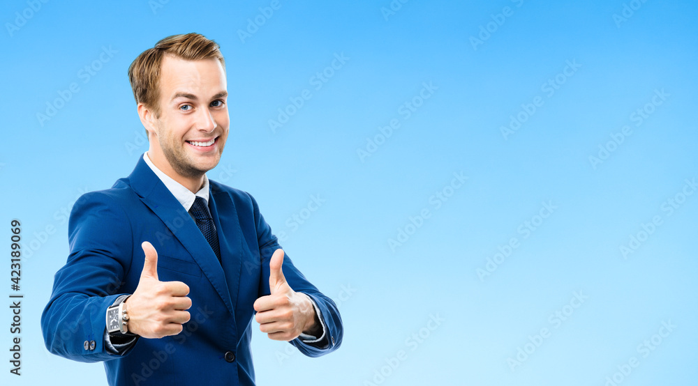 Fototapeta premium Portrait of confident businessman showing thumbs up hand sign gesture, on blue background. Business success concept. Young happy smiling man at studio.