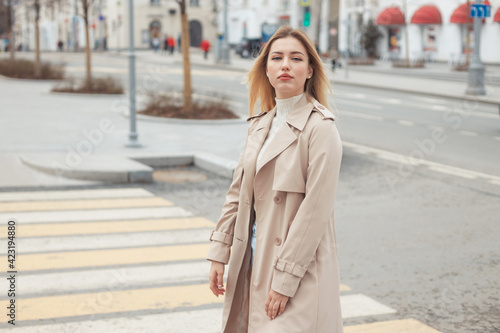 Young beautiful blonde hair woman in fashion clothes: beige trench coat, black boots and jeans crossing the road street. City lifestyle portrait.