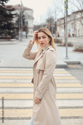 Young beautiful blonde hair woman in fashion clothes: beige trench coat, black boots and jeans crossing the road street. City lifestyle portrait. © Evgeniya Grande