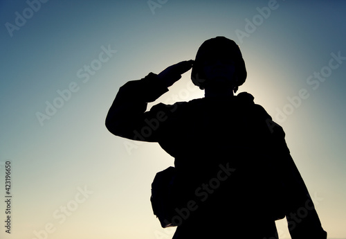 Silhouette of soldier in combat helmet and ammunition saluting on background of sunset sky. Army special forces fighter, Marines rifleman showing respect, greeting officer with salute gesture photo