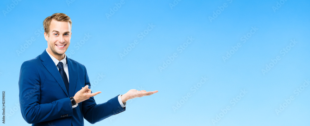 Portrait of confident businessman showing, giving or holding something, isolated on blue color background. Business success concept. Young happy smiling man at studio. Portrait of confident businessman showing, giving or holding something, isolated on blue color background. Business success concept. Young happy smiling man at studio.