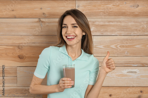 Young woman with glass of chocolate milk showing thumb up on wooden background