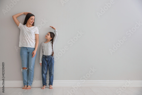 Little girl and mother measuring their height near light grey wall indoors. Space for text