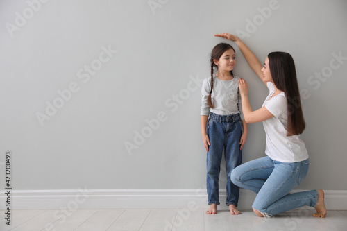 Mother measuring little girl's height near light grey wall indoors. Space for text photo
