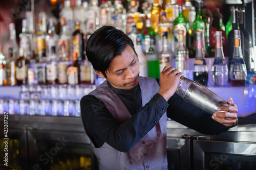 Smiling asian bartender shaking cocktail at the bar using metal mixer over colorful background with alcoholic beverages