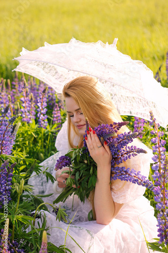 A gentle young long-haired girl in a white dress among a purple field  holding a lace umbrella and a bouquet of lupins. The concept of nature and romance.