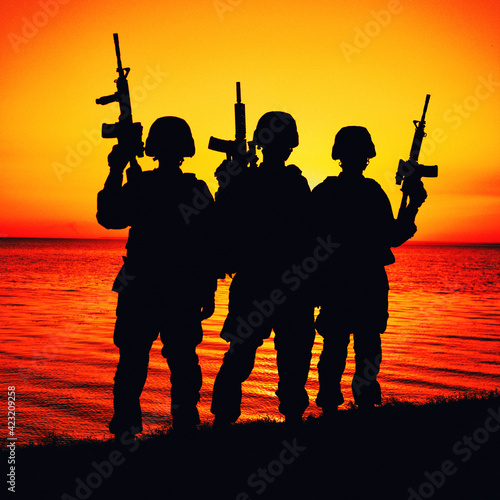 Silhouette of army special operations forces soldiers team, group of Marines or coast guard fighters crew in ammunition and combat helmets standing on seashore at sunset with raised assault rifles