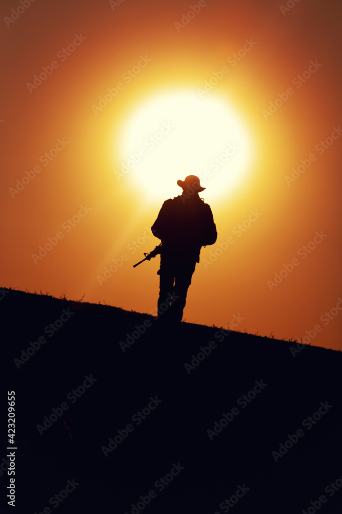 Silhouette of army special forces soldier armed with service rifle, commando, counter terrorist team fighter in boonie hat and uniform marching during mission, patrolling area on sunset or dawn time