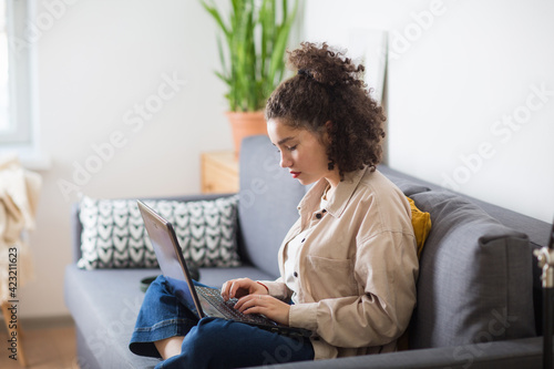 mixed rase girl is sitting on couch and looking at at computer screen laptop. e-learning at home.