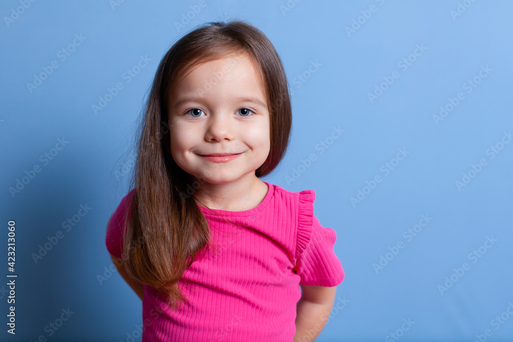A European girl 4-5 years old with blond hair blue eyes in a bright pink T-shirt on a blue background hides something behind her back. That's a nice smile. Emotions. A place for text or advertising. A European girl 4-5 years old with blond hair blue eyes in a bright pink T-shirt on a blue background hides something behind her back. That's a nice smile. Emotions. A place for text or advertising.