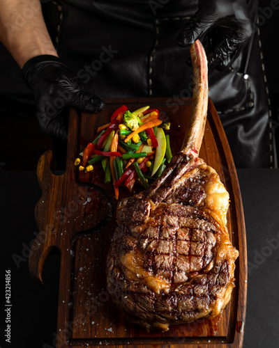 Hands holding cooked Tomahawk steak on a serving board. Low key image, vertical orientation
