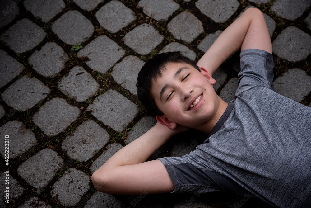 Casual outdoor spring portrait with copy space of a cute Asian_American tween boy wearing a gray t-shirt laying down on paver sidewalk looking relaxed and smiling at the camera Casual outdoor spring portrait with copy space of a cute Asian_American tween boy wearing a gray t-shirt laying down on paver sidewalk looking relaxed and smiling at the camera