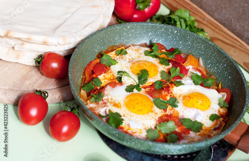 Traditional dish of israeli cuisine Shakshuka. Fried eggs with tomatoes, peppers, onions, cilantro and pita in a pan on light green background