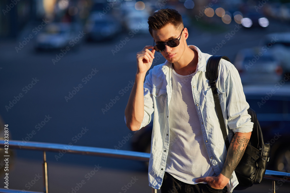 young man with a black backpack standing on a city street holding sunglasses in his hands.
