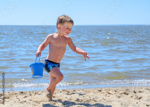 A boy, wet after swimming, smiles and runs with a toy bucket on the sand on the beach