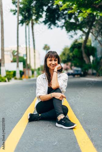 Urban vertical image of a woman sitting on the asphalt with her legs crossed and with her arm holding her face with a happy attitude