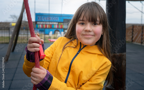 A child on the playground, a girl with long brown hair is played and smiles in a yellow jacket