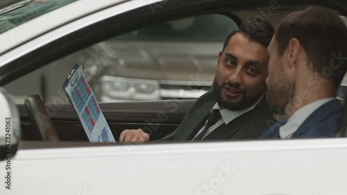 Medium close-up of two diverse colleagues wearing formal clothing, sitting in car, looking through documents, talking and working together photo