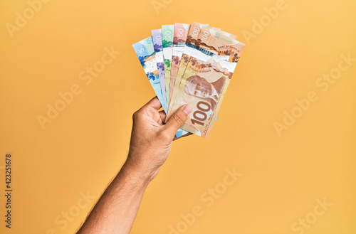 Hand of hispanic man holding canadian dollars over isolated yellow background. photo