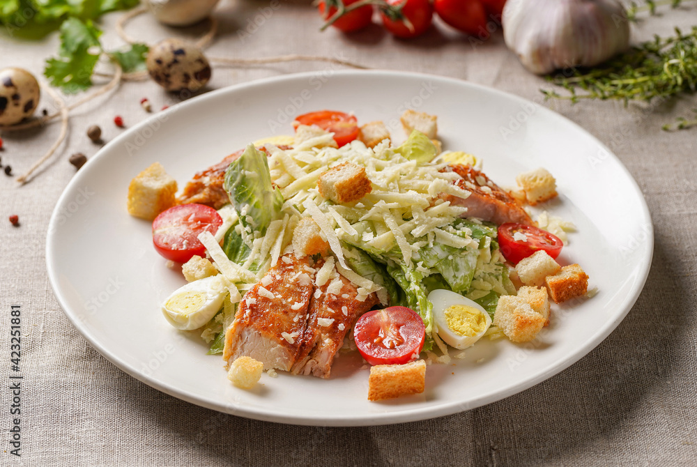 Caesar salad with chicken on a large white plate on the table covered with a light linen cloth on a beautiful background of vegetables and herbs. Beautiful bokeh. Caesar salad with chicken on a large white plate on the table covered with a light linen cloth on a beautiful background of vegetables and herbs. Beautiful bokeh.