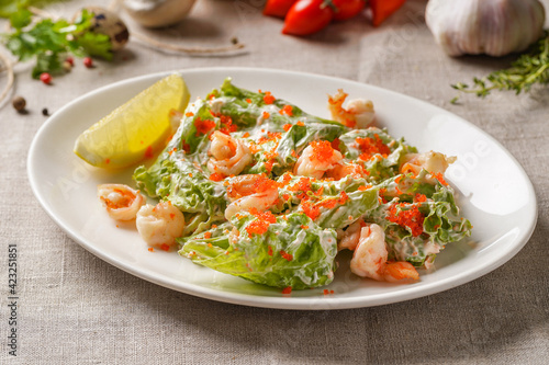 Salad with lettuce leaves, shrimp, tobiko caviar and sauce. With a slice of lemon on a plate. On the table covered with a light linen cloth on a beautiful background of vegetables and herbs.