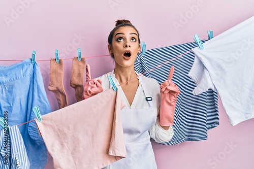 Beautiful brunette young woman washing clothes at clothesline amazed and surprised looking up and pointing with fingers and raised arms.