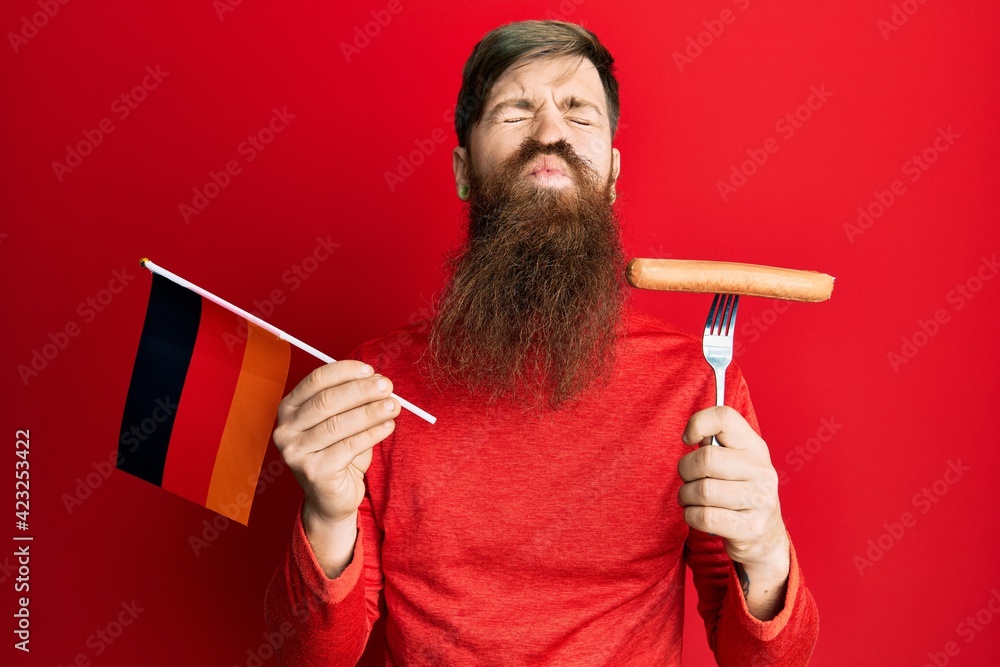 Fototapeta premium Redhead man with long beard holding fork with pork sausage and germany flag looking at the camera blowing a kiss being lovely and sexy. love expression.
