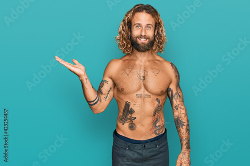 Handsome man with beard and long hair standing shirtless showing tattoos smiling cheerful presenting and pointing with palm of hand looking at the camera.