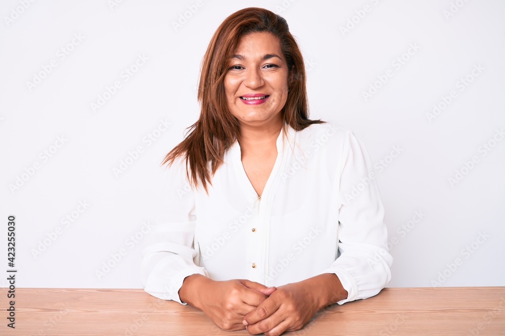 Middle age brunette hispanic business woman wearing casual white shirt sitting on the table with a happy and cool smile on face. lucky person. Middle age brunette hispanic business woman wearing casual white shirt sitting on the table with a happy and cool smile on face. lucky person.
