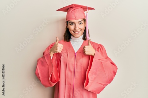 Young caucasian woman wearing graduation cap and ceremony robe success sign doing positive gesture with hand, thumbs up smiling and happy. cheerful expression and winner gesture.