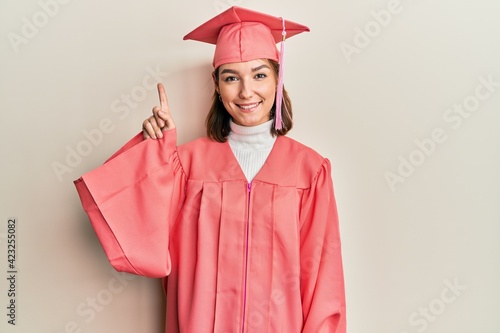 Young caucasian woman wearing graduation cap and ceremony robe showing and pointing up with finger number one while smiling confident and happy.