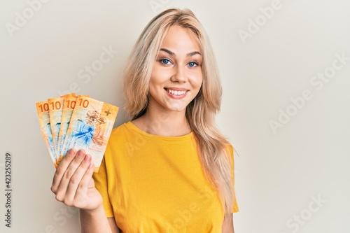 Beautiful caucasian blonde girl holding 10 swiss franc banknotes looking positive and happy standing and smiling with a confident smile showing teeth photo