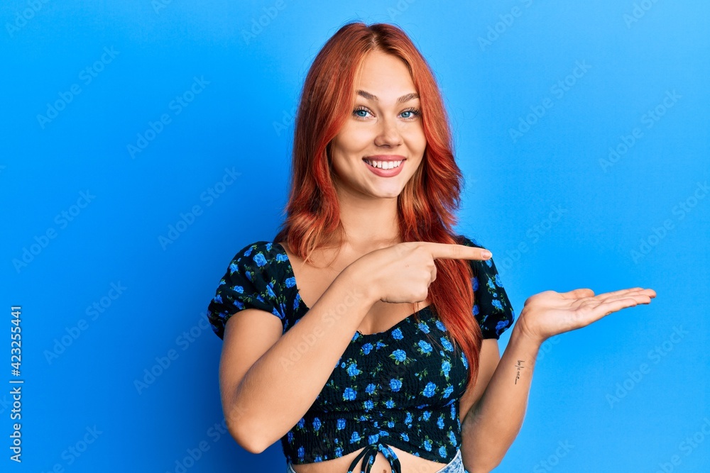 Young beautiful redhead woman wearing casual clothes over blue background amazed and smiling to the camera while presenting with hand and pointing with finger. Young beautiful redhead woman wearing casual clothes over blue background amazed and smiling to the camera while presenting with hand and pointing with finger.