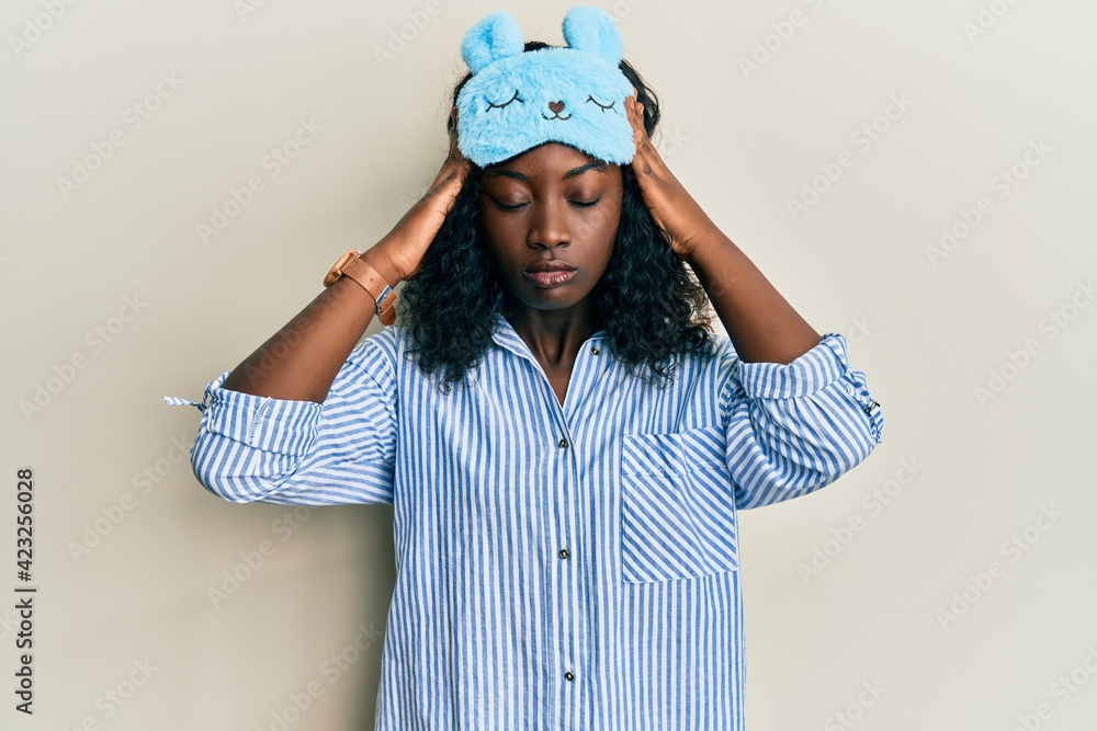 Beautiful african young woman wearing sleep mask and pajama suffering from headache desperate and stressed because pain and migraine. hands on head. Beautiful african young woman wearing sleep mask and pajama suffering from headache desperate and stressed because pain and migraine. hands on head.
