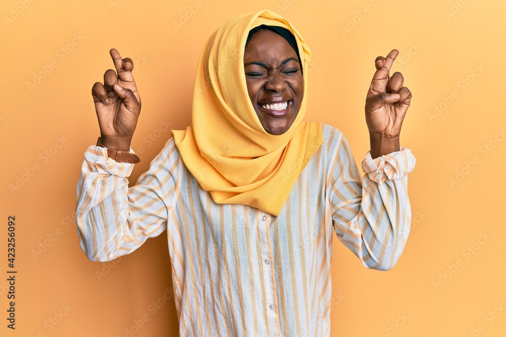 Beautiful african young woman wearing traditional islamic hijab scarf gesturing finger crossed smiling with hope and eyes closed. luck and superstitious concept. Beautiful african young woman wearing traditional islamic hijab scarf gesturing finger crossed smiling with hope and eyes closed. luck and superstitious concept.