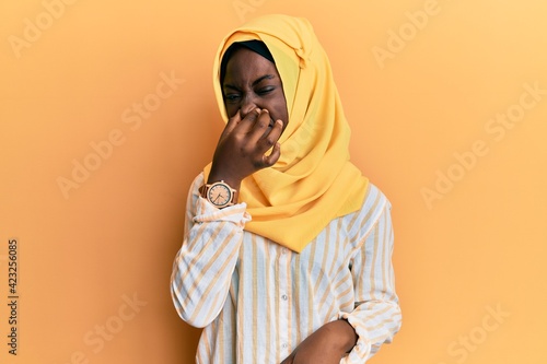 Beautiful african young woman wearing traditional islamic hijab scarf smelling something stinky and disgusting, intolerable smell, holding breath with fingers on nose. bad smell © Krakenimages.com
