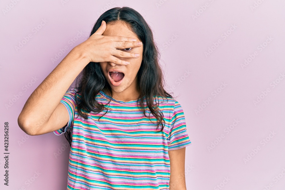 Hispanic teenager girl with dental braces wearing casual clothes peeking in shock covering face and eyes with hand, looking through fingers with embarrassed expression.