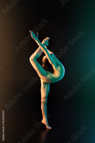 Young flexible girl isolated on a black studio background in neon light. A young female model is engaged in rhythmic gymnastics. Exercises for flexibility, balance Grace in movement, sport, and action