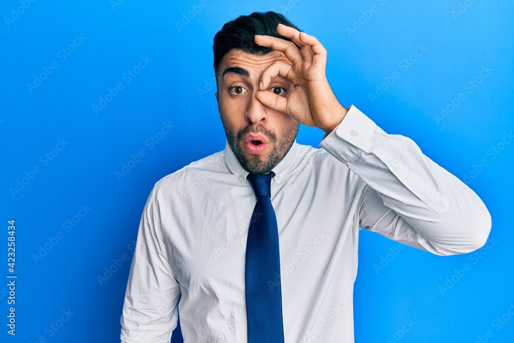 Young hispanic man wearing business clothes doing ok gesture shocked with surprised face, eye looking through fingers. unbelieving expression. Young hispanic man wearing business clothes doing ok gesture shocked with surprised face, eye looking through fingers. unbelieving expression.