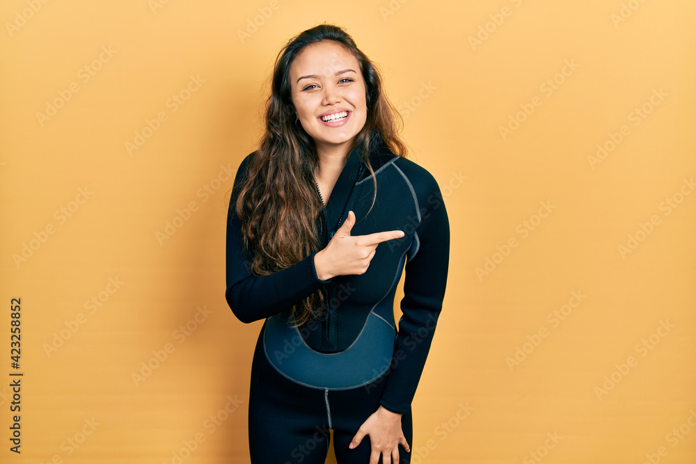 Young hispanic girl wearing diver neoprene uniform cheerful with a smile on face pointing with hand and finger up to the side with happy and natural expression Young hispanic girl wearing diver neoprene uniform cheerful with a smile on face pointing with hand and finger up to the side with happy and natural expression