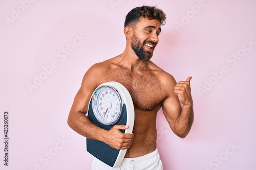 Young hispanic man standing shirtless holding weighing machine pointing thumb up to the side smiling happy with open mouth