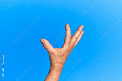 Hand of senior hispanic man over blue isolated background picking and taking invisible thing, holding object with fingers showing space