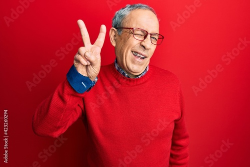 Handsome senior man with grey hair wearing casual clothes and glasses smiling with happy face winking at the camera doing victory sign. number two.