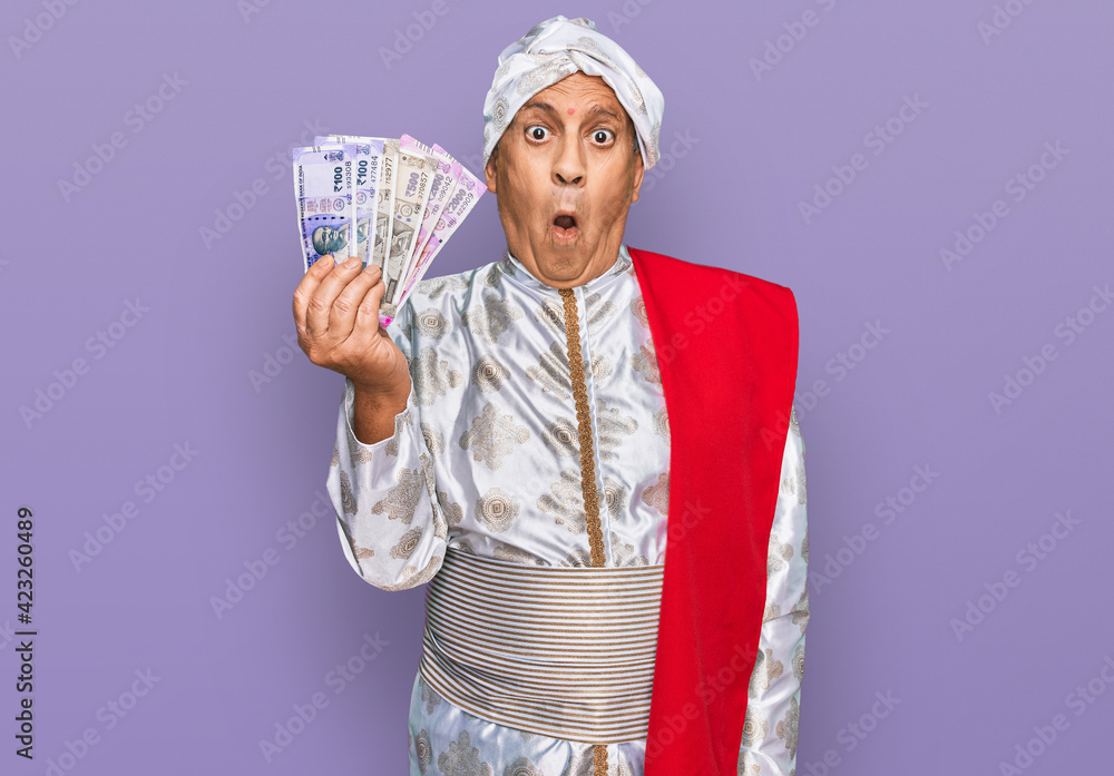 Senior hispanic man wearing sherwani costume holding indian rupee banknotes scared and amazed with open mouth for surprise, disbelief face Senior hispanic man wearing sherwani costume holding indian rupee banknotes scared and amazed with open mouth for surprise, disbelief face